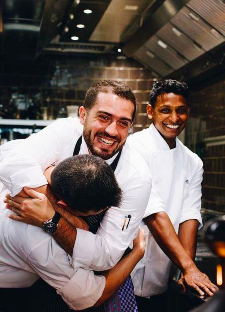 Tres cocineros con chaquetilla blanca riendo y abrazándose alegremente dentro de la cocina profesional del restaurante, mostrando un ambiente de trabajo dinámico y positivo.