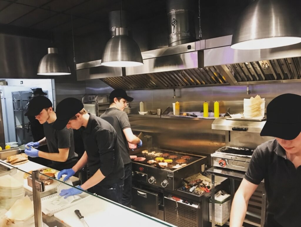Línea de cocina profesional con varios cocineros con gorra y guantes preparando hamburguesas en la zona de montaje y cocinando medallones de carne sobre una plancha caliente bajo campanas extractoras industriales.