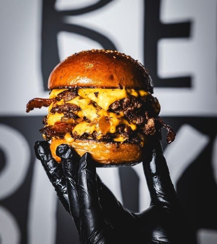 Primer plano de una hamburguesa con doble carne smash, abundante queso cheddar derretido y bacon, sostenida por unas manos con guantes de nitrilo negros frente a un fondo con letras difuminadas.