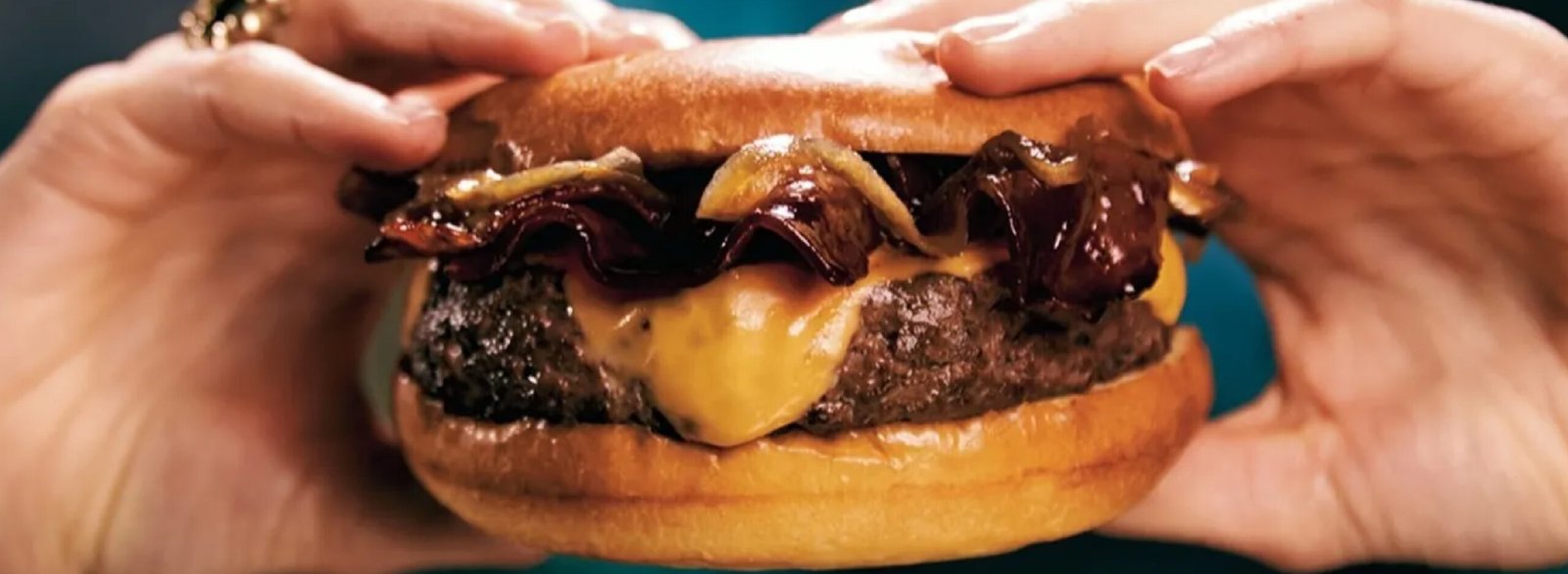 Primer plano horizontal de una hamburguesa gourmet sostenida por dos manos, mostrando una gruesa carne cocinada, queso cheddar fundido y tiras de bacon caramelizado crujiente sobre un fondo turquesa difuminado.