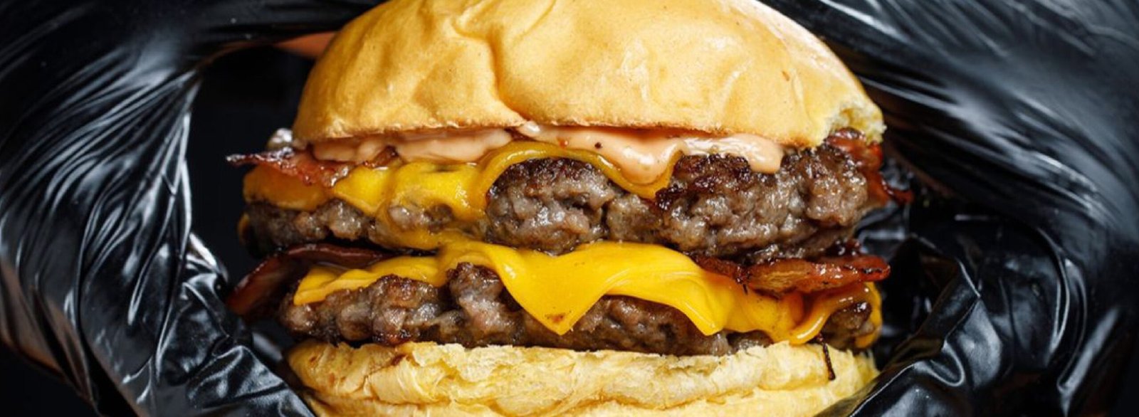 Primer plano horizontal de una hamburguesa con doble carne, queso cheddar fundido entre los medallones, bacon y salsa, enmarcada por unas manos con guantes negros de nitrilo.