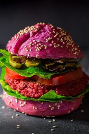 Hamburguesa vegana montada en un llamativo pan brioche de color morado con semillas de sésamo, que incluye un medallón de proteína vegetal, lechuga verde, rodajas de tomate y pepinillos sobre un fondo oscuro.