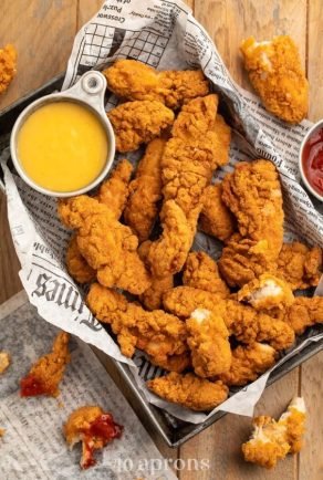 Bandeja metálica con papel de periódico que contiene tiras de pollo deshuesado con un rebozado grueso muy crujiente y dorado, acompañadas de cuencos con salsa de mostaza y kétchup sobre una mesa de madera.